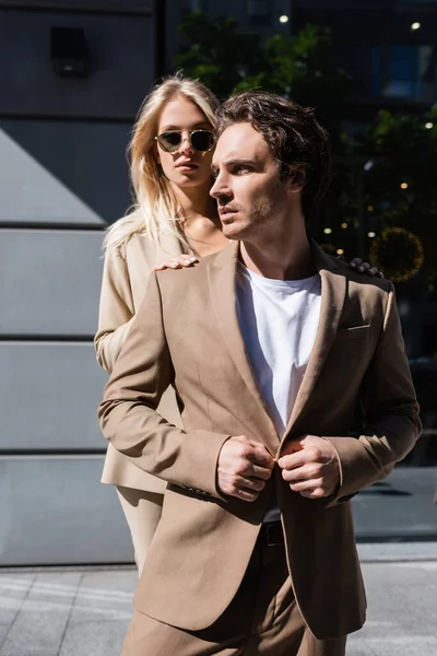 Young man buttoning beige blazer and looking away near stylish woman in sunglasses — Stock Photo