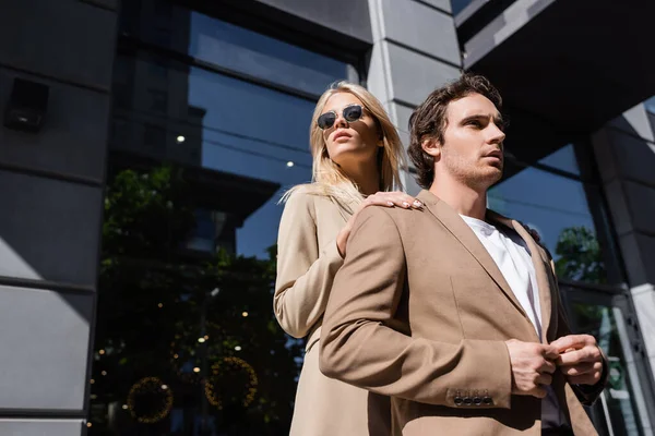 Low angle view of blonde woman in sunglasses touching shoulders of man buttoning stylish blazer — Stock Photo