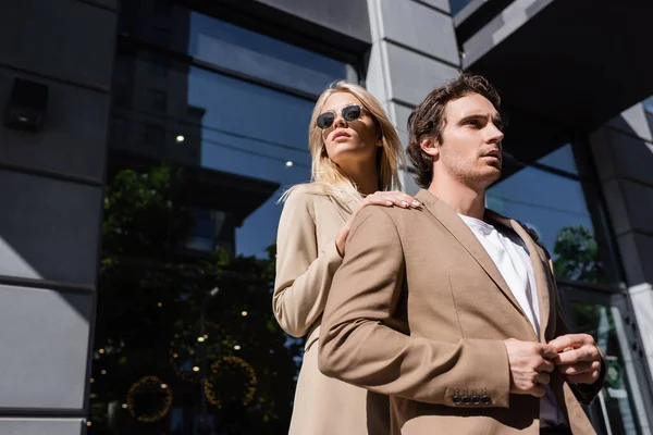 Vista de ángulo bajo de mujer rubia en gafas de sol tocando hombros de hombre abotonando elegante chaqueta - foto de stock