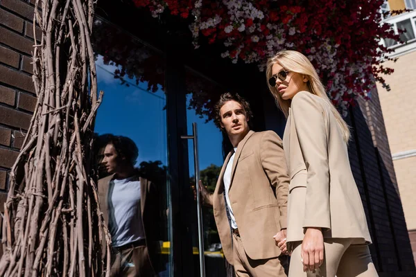 Hombre de moda abriendo puertas de vidrio cerca de mujer rubia en gafas de sol - foto de stock