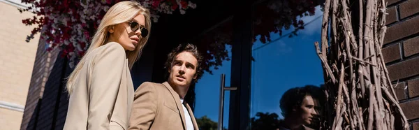 Low angle view of brunette man looking at woman in stylish sunglasses near glass doors, banner — Stock Photo