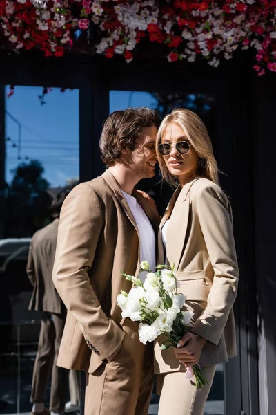 Sonriente hombre de pie con la mano en el bolsillo cerca de la mujer con estilo con flores blancas - foto de stock