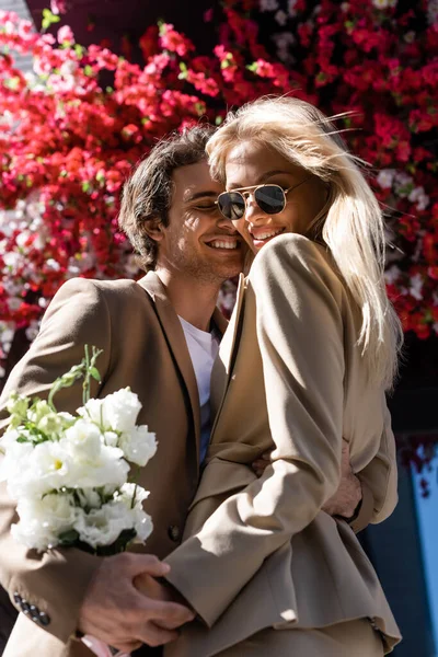 Mujer complacida en gafas de sol sosteniendo ramo cerca de hombre feliz abrazándola al aire libre - foto de stock