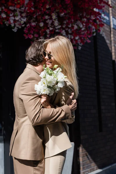 Vue latérale du jeune homme embrassant femme dans des lunettes de soleil tenant bouquet sur la rue de la ville — Photo de stock