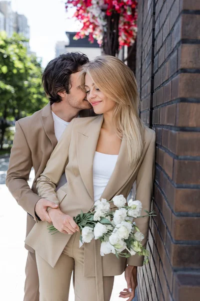 Brunette man kissing happy and stylish woman holding white flowers on street — Stock Photo