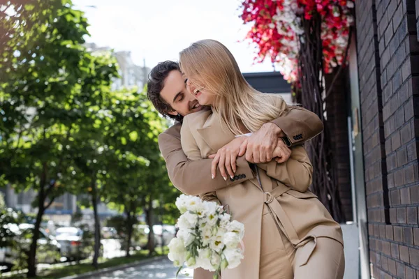 Happy man embracing blonde woman holding white flowers on city street — Stock Photo