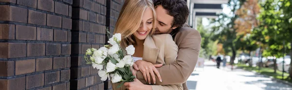 Young man embracing cheerful blonde woman holding flowers near brick wall, banner — Stock Photo