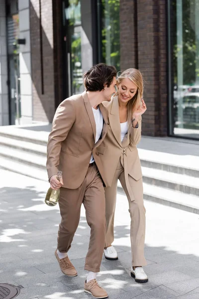 Full length of young man with champagne bottle walking near blonde woman on street — Stock Photo