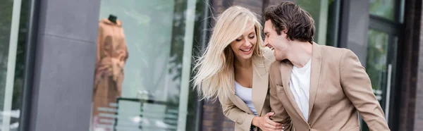 Cheerful young couple in beige blazers laughing on city street, banner — Stock Photo
