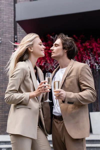 Couple heureux en costumes à la mode tenant des verres de champagne et se regardant dans la rue — Photo de stock