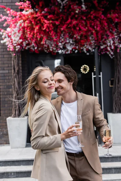 Donna gioiosa con gli occhi chiusi e sorridente uomo bruna che tiene bicchieri di champagne per strada — Foto stock