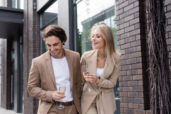 Alegre pareja joven en trajes beige caminando con copas de champán en la calle de la ciudad - foto de stock