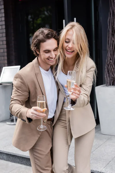 Casal alegre em ternos elegantes bege segurando copos de champanhe na rua na cidade — Fotografia de Stock