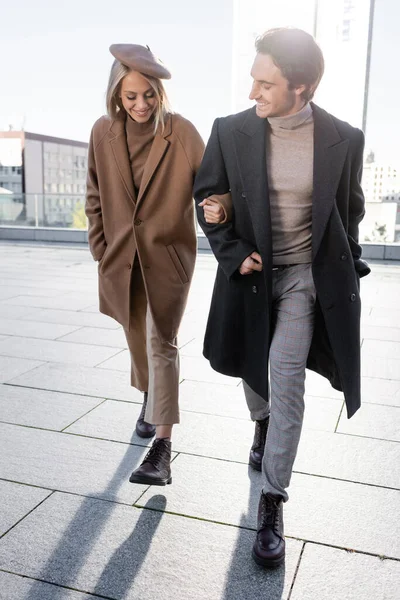 Pleine longueur de couple heureux avec les mains dans des poches de manteaux marchant en ville — Photo de stock