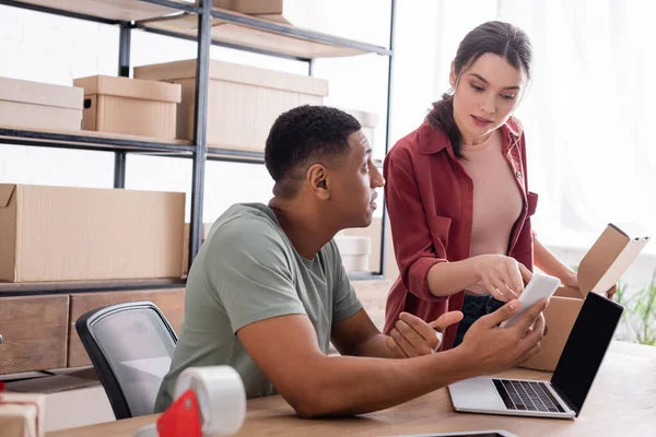 Afrikanischer Verkäufer hält Smartphone neben Kollegin mit Karton im Geschäft — Stockfoto