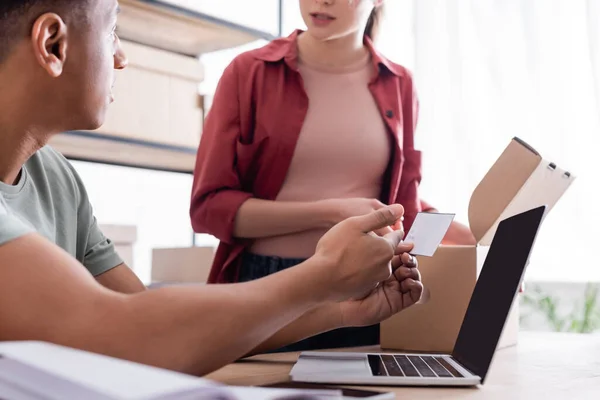 Vendedor afroamericano sosteniendo etiqueta de envío cerca de la computadora portátil y colega con caja de cartón en la tienda - foto de stock