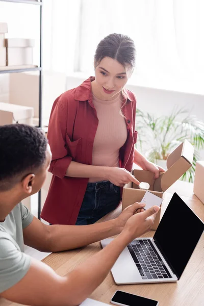 Young seller pointing at carton box near african american colleague with shipping label and devices in store — Stock Photo