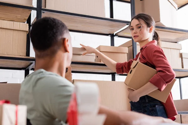 Young seller holding carton box near blurred african american colleague in online web store — Stock Photo