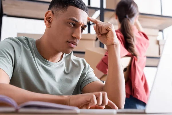 Venditore afroamericano utilizzando laptop offuscata vicino collega e pacchetti nel negozio online web — Foto stock