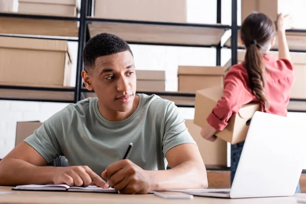 Propietario de una tienda web afroamericana en línea escribiendo en un cuaderno cerca de dispositivos y colega con paquetes - foto de stock