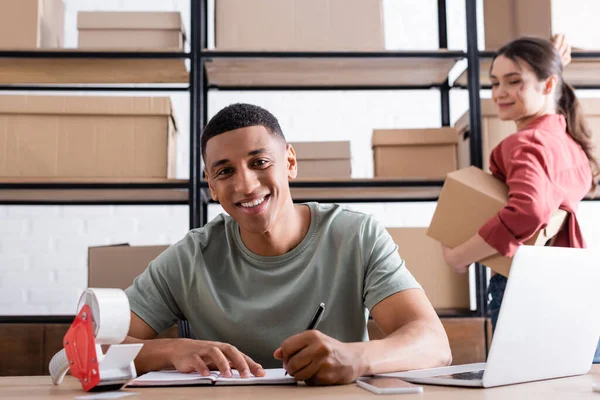 Sonriente afroamericano dueño de la tienda web en línea escribiendo en el cuaderno cerca de colega borrosa con cajas - foto de stock