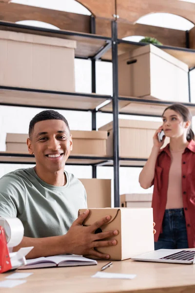 Propietario afroamericano positivo que sostiene la caja de cartón cerca del ordenador portátil y colega borroso en la tienda web en línea - foto de stock