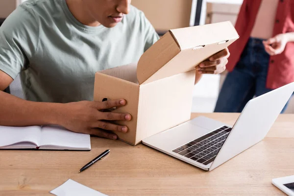 Vista recortada del vendedor afroamericano que sostiene la caja de cartón cerca del ordenador portátil en la tienda web en línea - foto de stock