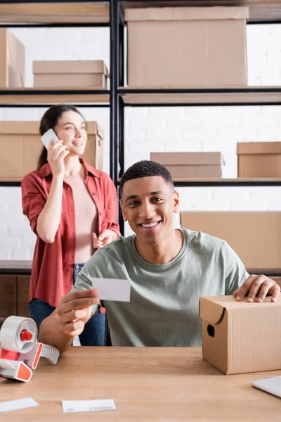 Alegre vendedor afroamericano con tarjeta de visita y caja de cartón mientras su colega habla en el teléfono inteligente en la tienda - foto de stock