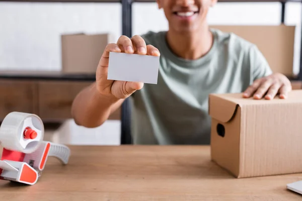 Vista recortada de la borrosa tarjeta de visita del vendedor afroamericano sosteniendo cerca de caja de cartón y cinta adhesiva en la tienda - foto de stock