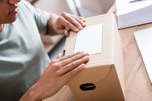 Vista recortada del vendedor afroamericano aplicando etiqueta de envío en caja de cartón en tienda - foto de stock