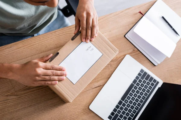 Cropped view of african american seller holding cardboard box with shipping label near laptop and notebook in shop — Stock Photo