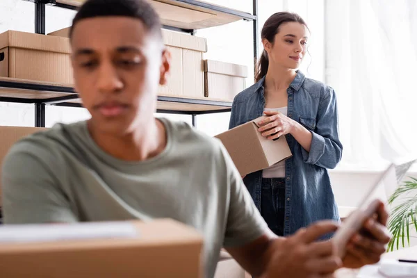 Joven vendedor sosteniendo caja de cartón cerca borrosa africana colega americano con tableta digital en la tienda web en línea - foto de stock