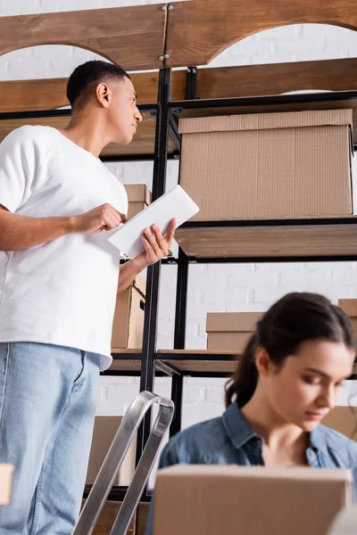 African american seller using digital tablet near carton boxes and blurred colleague in online web store — Stock Photo