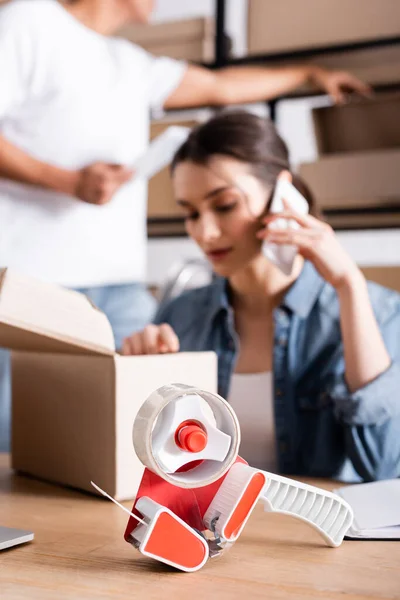 Adhesive tape near blurred seller talking on smartphone and carton box in store — Stock Photo