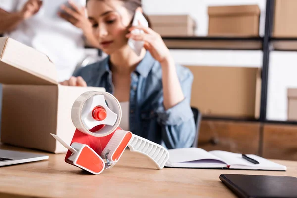 Adhesive tape near notebooks and blurred seller talking on smartphone near package in store — Stock Photo