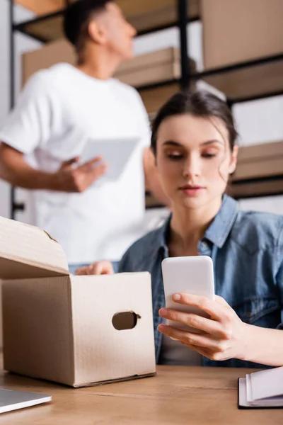 Blurred seller holding smartphone near cardboard box and blurred african american colleague in online web store — Stock Photo