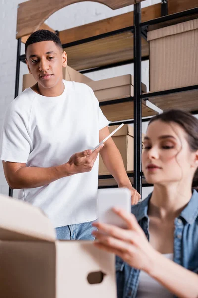 Vendedor afroamericano sosteniendo tableta digital cerca de colega borrosa con teléfono inteligente y caja de cartón en la tienda - foto de stock