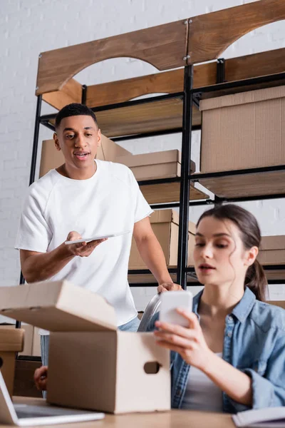 African american seller holding digital tablet near blurred colleague with cellphone and cardboard boxes in online web store — Stock Photo