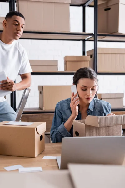 Vendedor hablando en teléfono inteligente cerca de cajas de cartón y colega afroamericano con tableta digital en la tienda - foto de stock