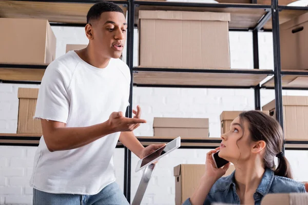 African american seller with digital tablet talking to colleague talking on smartphone in online web store — Stock Photo