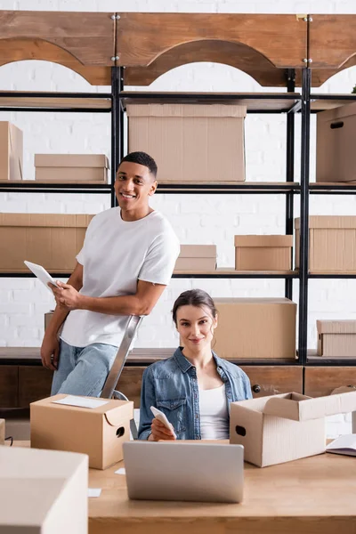 Smiling interracial sellers with devices looking at camera near carton boxes in online web store — Stock Photo