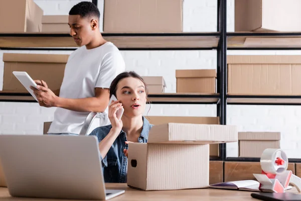 Seller talking on smartphone near cardboard boxes and african american colleague with digital tablet in online web store — Stock Photo