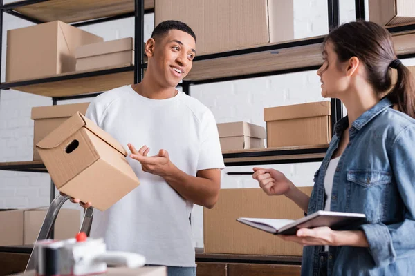 African american seller pointing at cardboard box near colleague with notebook in online web store — Stock Photo