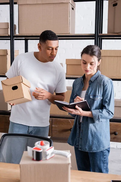 Vendeurs multiethniques écrivant sur ordinateur portable près de boîtes en carton dans une boutique en ligne — Photo de stock