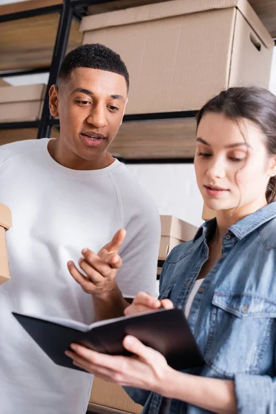 Afrikanischer Verkäufer zeigt in Online-Webshop auf Notizbuch neben verschwommenem Kollegen — Stockfoto