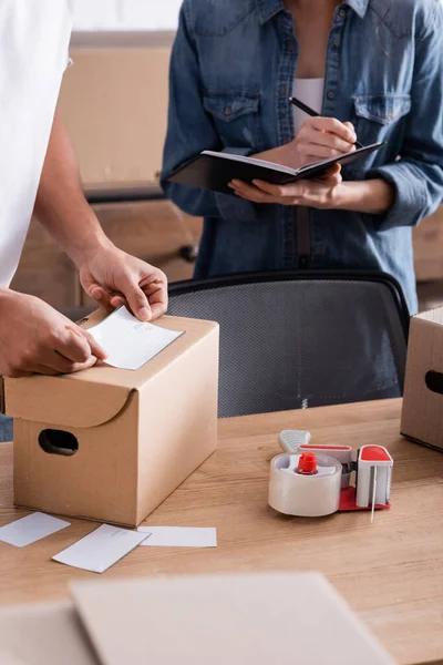 Vista recortada del vendedor afroamericano aplicando la etiqueta de envío en la caja de cartón cerca de colega escribiendo en el cuaderno en la tienda web en línea - foto de stock