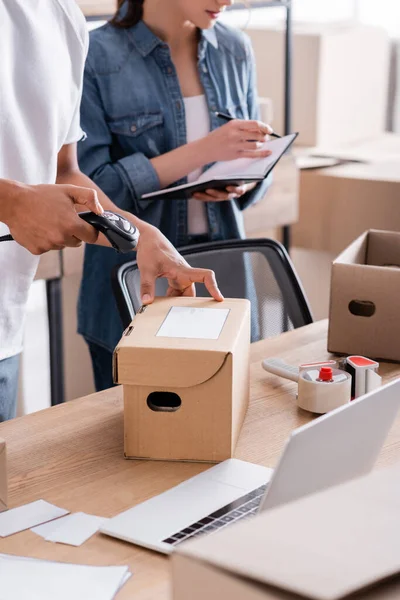 Ausgeschnittene Ansicht eines afrikanisch-amerikanischen Verkäufers, der Pakete in der Nähe von Laptop und Kollegen im Online-Webshop scannt — Stockfoto