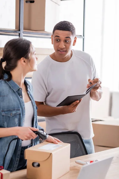 Afrikanischer Verkäufer hält Notizbuch in der Nähe des Kollegen, der Paket scannt — Stockfoto