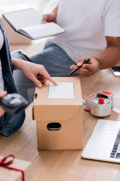 Vue recadrée du vendeur pointant vers l'étiquette d'expédition sur la boîte en carton près d'un collègue afro-américain avec ordinateur portable en magasin — Photo de stock