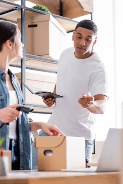 African american seller holding notebook near colleague with scanner and carton box in online web store — Stock Photo